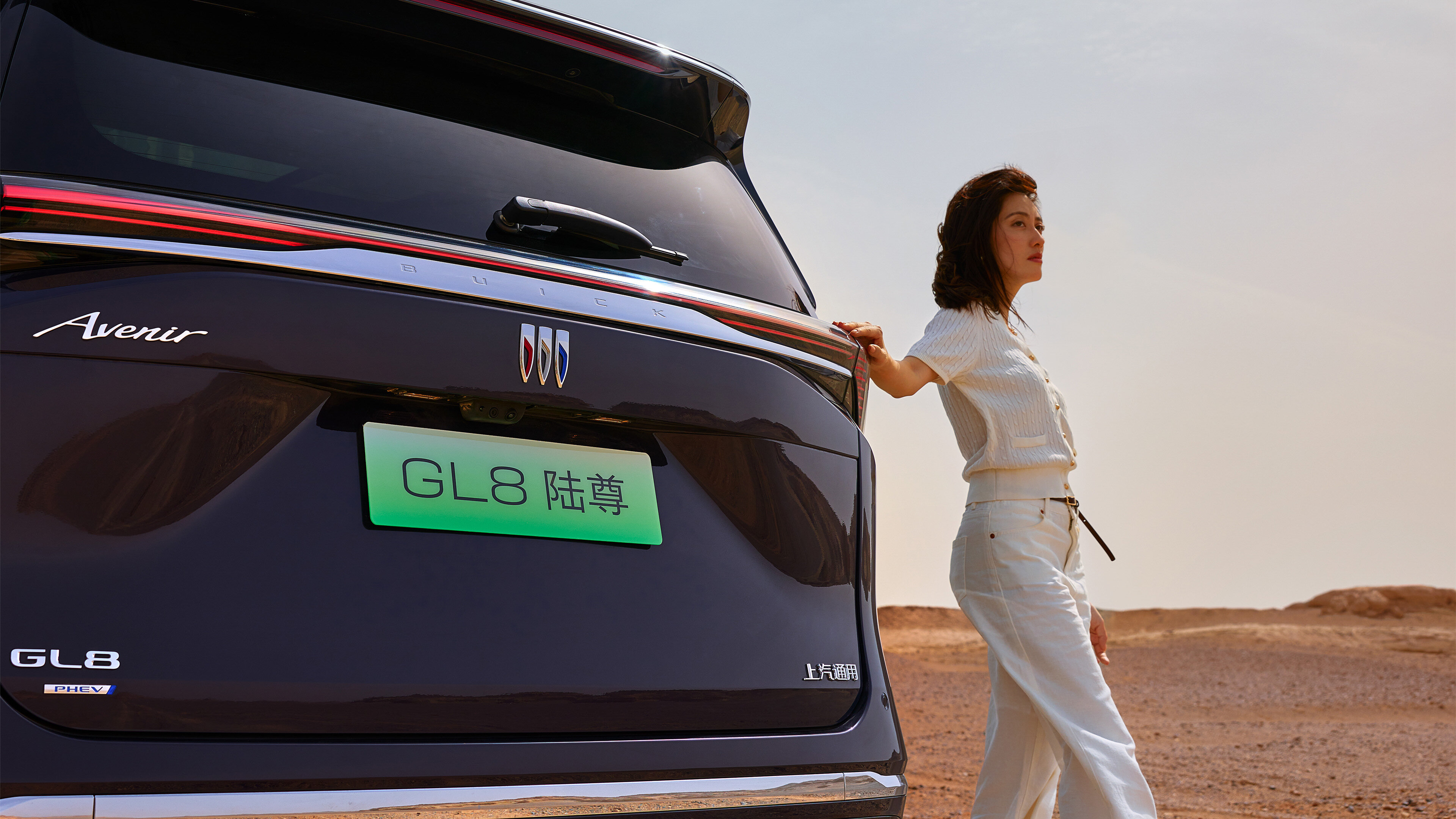 Rear three-quarter view of 2026 Buick GL8 Lushang in Obsidian Black, desert backdrop, woman leaning against tailgate