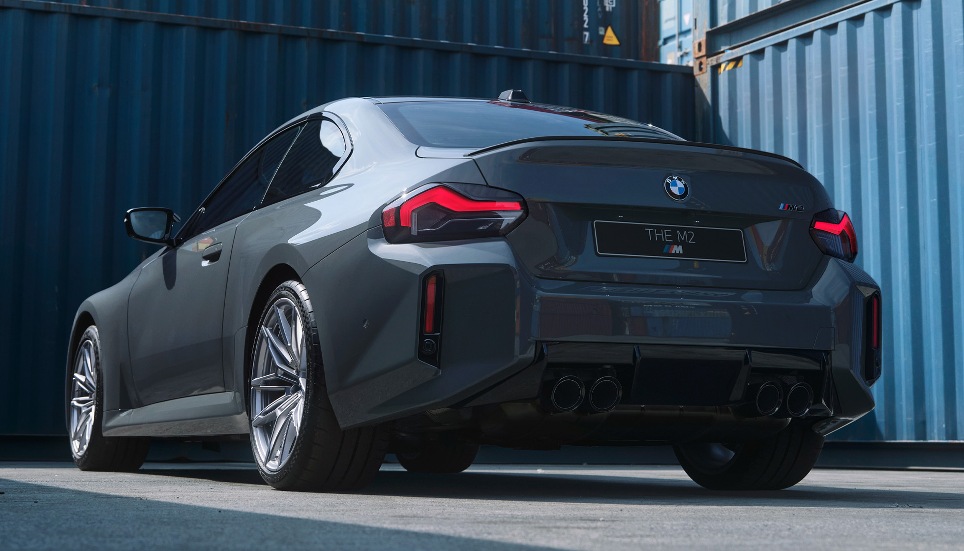 Rear three-quarter view of the Brilliance BMW BMW M2 2024 model in dark gray, against a blue shipping container background.