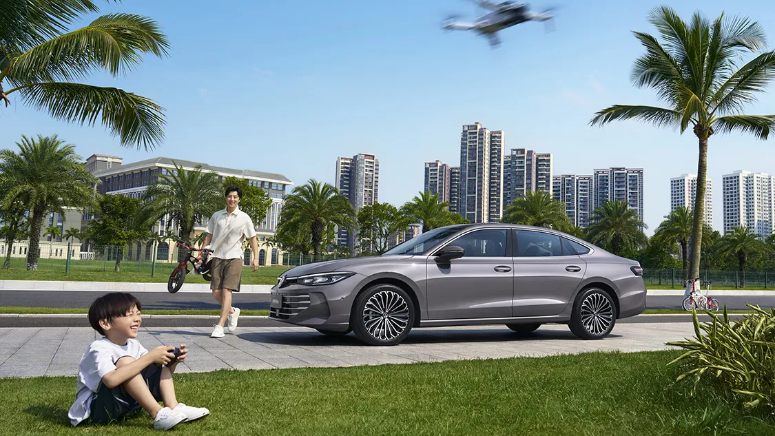 Vue latérale gauche de la Passat 2026 de SAIC Volkswagen, carrosserie gris « Ciel galactique », stationnée près d’un parc urbain verdoyant, immeubles et palmiers en arrière-plan